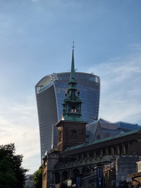 A detailed view of a historic church with a tall, green spire topped by a weather vane, set against the modern, curved glass skyscraper known as the Walkie Talkie in London. The church's dark brickwork features arched windows and ornamental stonework, with a weathered roof and lush green trees partially obscuring the lower part of the structure. The background displays the sky during late afternoon or early evening, with soft light illuminating the scene. The juxtaposition of old and new architectural styles reflects the city's blend of historical and contemporary elements. The outdoor setting shows a well-maintained area likely served by professional gardening and landscaping services, such as those offered by Gardeners Twickenham, highlighting a combination of classic and modern urban landscape features.