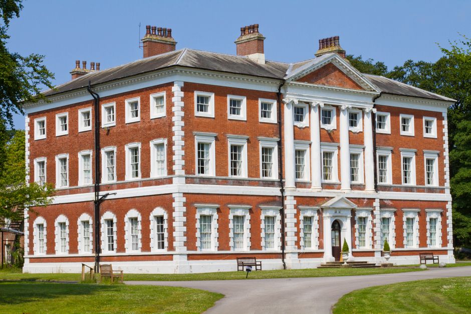 A historic red brick building with white stone detailing, featuring a symmetrical facade with multiple sash windows and a central entrance with classical columns, situated in a well-maintained garden. The garden displays trimmed lawns, a paved pathway, and neatly manicured shrubs, with mature trees in the background providing shade. The sky is clear and blue, highlighting the crisp textures of the stonework and the lush greenery, as Gardeners Twickenham is performing landscape or lawn maintenance around the stately property, with evidence of trimming or careful garden upkeep indicative of professional landscaping work around York House gardens.